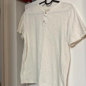 Vit kortärmad t-shirt från H&M - Säljer en basic vit t-shirt från H&M i ekologisk bomull. T-shirten har korta ärmar, rund halsringning och två knappar framtill som ger en chill look. Perfekt till jeans eller shorts för en enkel och clean stil. Använd 2-3 gånger 