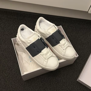 Valentino Open sneakers vit/navy skinn - Snygga Valentino Open sneakers i vitt skinn med bred svart rem över vristen och diskret guldlogga på plösen. Klassisk rund tå, platt sula och coola nitar baktill. Perfekta för dig som gillar stilrena och lyxiga sneakers med edge.