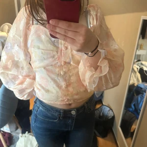 Blommig croppad blus med puffärm - Superfin blommig blus i ljusa pastellfärger med croppad passform och stora puffärmar. Toppen har knappar framtill och volangdetaljer vid ärmsluten. Perfekt för dig som gillar romantisk och trendig stil.