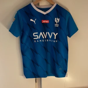 Al Hilal Neymar Jr #10 matchställ Puma - Säljer ett blått Al Hilal matchställ med Neymar Jr #10. Tröjan har korta ärmar, vit krage och coolt mönster, med tryck från Savvy Games Group och Jahez. Shortsen är blå med klubbmärke och nummer 10. Perfekt för dig som älskar fotboll och vill sticka ut på planen.