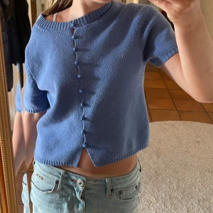 Blå stickad kofta från Massimo Dutti - Supersöt blå stickad kofta från Massimo Dutti med kort ärm och rund hals. Koftan har dekorativa knappar framtill i samma färg och en croppad passform. Perfekt till jeans eller kjol för en chill och trendig look. Passar till alla årstider!