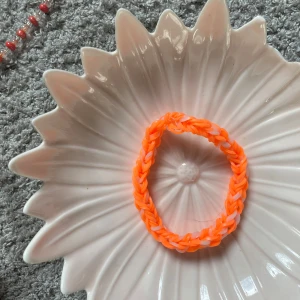 Orange och vitt armband i gummiband - Säljer ett handgjort armband i orange och vitt, flätat av elastiska gummiband. Perfekt accessoar för att addera lite färg till din stil. Passar de flesta handleder tack vare det stretchiga materialet.