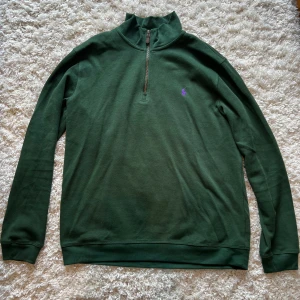 Grön halvzip tröja från Ralph Lauren - Mörkgrön långärmad tröja från Ralph Lauren med halv dragkedja och lila broderad logga på bröstet. Tröjan har ribbade muddar och hög krage, perfekt för lager-på-lager-stil.