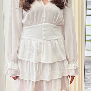 Vit volangklänning med knappar - Superfin vit klänning med volanger och markerad midja. Klänningen har långa ärmar med breda manschetter, v-ringning och knappar framtill. Väldigt fint skick💗