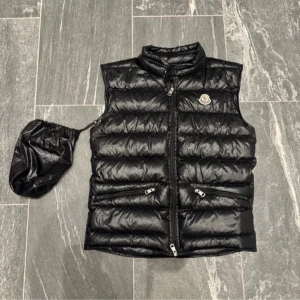 Svart dunväst från Moncler - Snygg svart dunväst från Moncler med hög krage och dragkedja framtill. Västen har två snedställda fickor med dragkedja och Moncler-logga på bröstet. Levereras med tillhörande förvaringspåse, perfekt att ta med på språng.