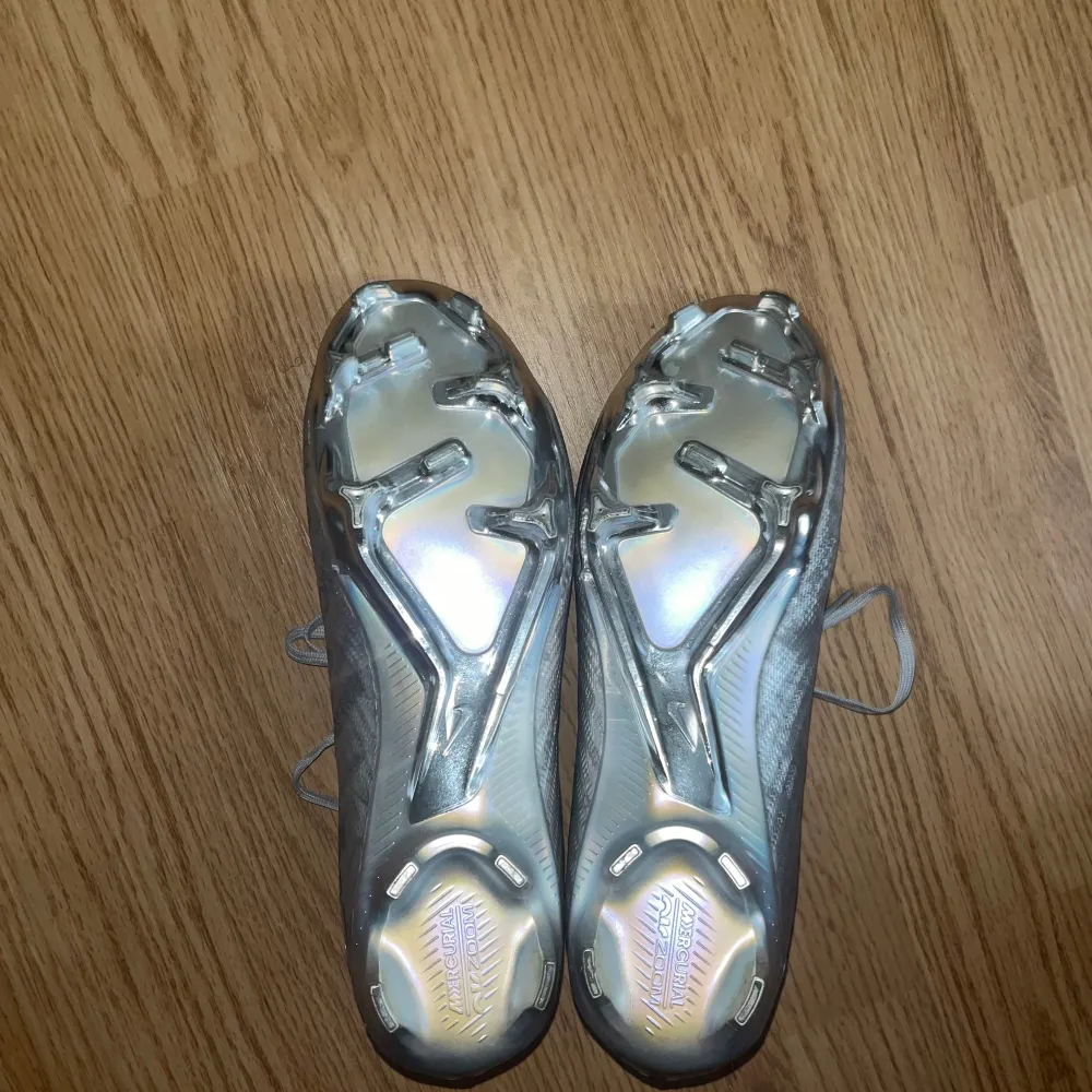 Säljer ett par Nike Mercurial fotbollsskor i metallic silver med guldigt märke på sidan. Skorna har snörning och mönstrad ovandel med textdetaljer. Yttersulan är i blankt silver med fasta dobbar, perfekt för spel på gräs och konstgräs!. Kengät.
