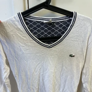 Vit v-ringad tröja från Lacoste - Säljer en vit långärmad tröja från Lacoste med v-ringning och mörkblå/vita ränder vid halsen. Tröjan har diskret rutmönster och en broderad Lacoste-logga på bröstet. Baksidan har ett tydligt rutigt mönster i mörkblått och vitt.