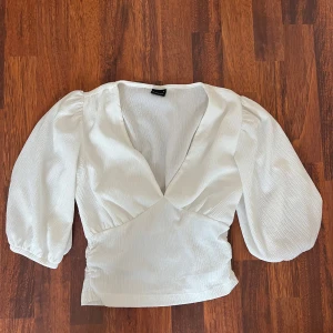 Vit omlottopp från Gina Tricot - Säljer en vit omlottopp från Gina Tricot med trekvartsärmar och markerad midja. Toppen har v-ringning och lätt puffiga ärmar som ger en trendig look. Perfekt att styla med jeans eller kjol! Aldrig använd och priset kan diskuteras 