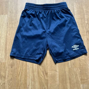 Mörkblå shorts från Umbro - Mörkblå sportiga shorts från Umbro med elastisk midja och logga på ena benet. Perfekta för träning eller fotboll. Klassisk design utan extra detaljer.