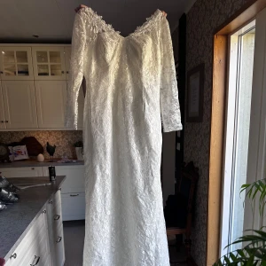 Vit längklänning/ bröllopsklänning i spets med knappar - Elegant vit långklänning i spets med v-ringning och långa ärmar. Klänningen har fina knappar längs ryggen och ett vackert blommigt spetsmönster över hela plagget. Perfekt för speciella tillfällen.