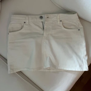 Vit jeanskjol - Säljer en vit kort jeanskjol med klassisk femficksdesign och knapp- samt dragkedjestängning framtill. Kjolen har raka linjer och är perfekt för en stilren look. Passar till många olika outfits och är enkel att matcha.