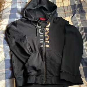 Svart hoodie med dragkedja från Hugo boss  - Säljer en svart hoodie från Hugo med dragkedja och huva. Tröjan har stora tryckta bokstäver i guld och silver längs dragkedjan samt fickor framtill. 