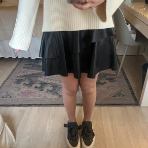 Svart volangkjol i skinnmaterial - Säljer en svart kortkjol med volangdetaljer. Kjolen har en lätt glansig finish och är tillverkad i ett syntetmaterial. Perfekt för dig som gillar en trendig och stilren look.🖤