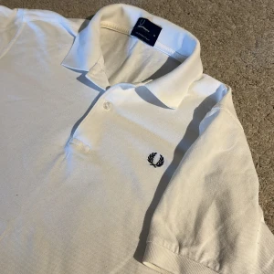 Fred Perry Pike  - Klassisk vit pikétröja från Fred Perry med broderad logga på bröstet. Tröjan har krage, knappar vid halsen och korta ärmar. Perfekt för en stilren och sportig look.