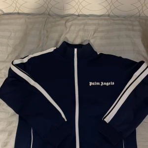 Mörkblå track jacket från Palm Angels - Säljer en mörkblå track jacket från Palm Angels med vita ränder längs ärmarna och dragkedja framtill. Jackan har hög krage och logga broderad på bröstet. Perfekt för en sportig och trendig look. Helt ny aldrig använd 