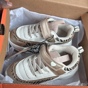 Vita och beige sneakers med zebramönster från Zigzag - Säljer ett par snygga sneakers från Zigzag i vitt och beige med zebramönstrade detaljer och ett litet hjärta på sidan. Skorna har både snörning och kardborreband för enkel påtagning. Perfekta för dig som har små barn som behöver skor. HELT NYA!