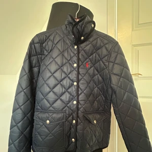 Mörkblå  quiltad jacka från Polo Ralph Lauren - Snygg svart quiltad jacka från Polo Ralph Lauren med hög krage och tryckknappar. Jackan har två fickor framtill och diskret logga broderad på bröstet. Perfekt för kyligare dagar och ger en stilren look.