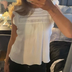 Vit blus med spetsdetaljer - Söt vit blus med spetsdetaljer upptill och volang vid axlarna. Blusen är ärmlös och har en luftig passform, perfekt för varmare dagar.
