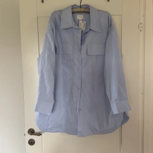 Ljusblå oversized skjorta från H&M - Säljer en ljusblå oversized skjorta från H&M med klassisk krage och knäppning framtill. Skjortan har två bröstfickor och långa ärmar med manschett. Perfekt för en avslappnad och trendig look. Tunt foder. 