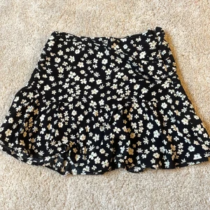 Svart blommig kjol med volang - Supersöt svart kjol med vita blommor och volang nertill. Kjolen är kort i längden. Använt mycket under sommaren men jättebra skick.  Storlek: 34/32 pris går att diskutera