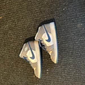 Nike Air Jordans Mid 1 True Blue - Säljer ett par Nike Air Jordans med grå och blå färgkombination. Skorna har klassisk hög modell med blå swoosh och snörning. Perfekta för dig som gillar streetwear och vill ha en ikonisk sneaker i garderoben.