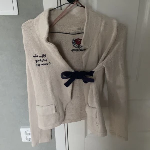 Stickad jacka med rosett från Odd Molly - Krämvit stickad jacka från Odd Molly med broderad text och ros på bröstet och ryggen. Jackan har lång ärm, två fickor fram och knyts med en mörkblå rosett. Snygg detalj med broderi och dekorativ knappslå bak.