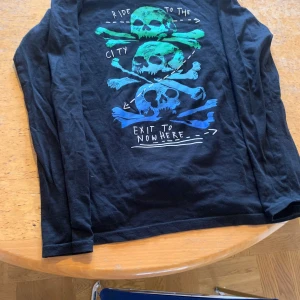 Svart långärmad tröja med döskalletryck från Lindex - Cool svart långärmad tröja från Lindex med färgglatt döskalletryck i grönt och blått på framsidan. Texten 'Ride to the city, exit to nowhere' ger en edgy vibe. Perfekt för dig som gillar streetstyle och grafiska prints.