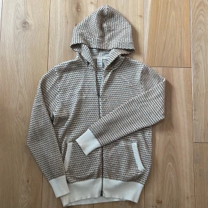 Beige och vit stickad kofta med dragkedja - Säljer en stickad cardigan i beige och vitt med småprickigt mönster. Den har dragkedja framtill, två fickor och ribbade muddar vid ärmslut och nederkant. Perfekt för lager-på-lager och chill vardagsstil.