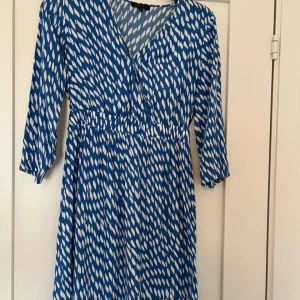 Blåvit mönstrad klänning från Caroll - Säljer en blå klänning från Caroll med vitt abstrakt mönster. Klänningen har v-ringning, resår i midjan och trekvartsärm. Perfekt för vår och sommar, lätt och luftig modell.