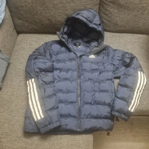 Adidas pufferjacka med huva - Säljer en blå pufferjacka från Adidas med huva och dragkedja. Jackan har de klassiska vita Adidas-ränderna längs ärmarna och logga på bröstet. Perfekt för kyligare dagar och riktigt skön att ha på sig. Ny kvalitet med ingen problem förutom en liten prick skada på ena armbågen.