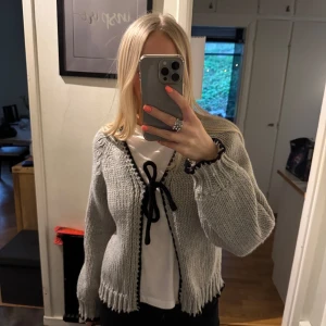 Grå stickad kofta med svarta detaljer - Säljer en mysig grå stickad kofta med svarta virkade kanter och knytband framtill. Koftan har ballongärmar och ribbade muddar. Perfekt att slänga över en t-shirt för en chill look.