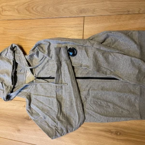 Grå hoodie med dragkedja C.P Company  - Säljer en grå hoodie från C.P Company med dragkedja framtill och huva. Tröjan har fickor på sidorna och en liten logga på ärmen. Perfekt för en avslappnad stil.