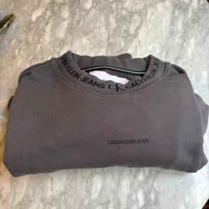 Grå långärmad tröja från Calvin Klein Jeans - Säljer en stilren grå långärmad tröja från Calvin Klein Jeans. Tröjan har rund halsringning med logotypdetaljer runt kragen och diskret tryck på bröstet. Perfekt för en clean och avslappnad look.