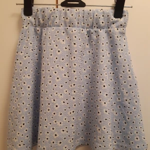 Ljusblå blommig kjol, strlk 130 - Supersöt ljusblå kjol från Lager 157 med små vita blommor och svarta prickar. Kjolen har resår i midjan och är gjord i ett mjukt, strukturerat tyg. Perfekt för dig som gillar en lekfull och somrig stil!