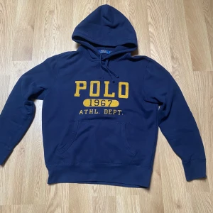 Mörkblå hoodie från Polo Ralph Lauren - Säljer en mörkblå hoodie från Polo Ralph Lauren med gul text och tryck framtill. Klassisk känguruficka och justerbar huva. Perfekt för en sportig och avslappnad stil.