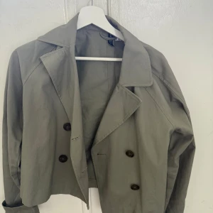 Grön croppad trenchcoat från H&M Divided - Snygg grå croppad trenchcoat från H&M Divided med dubbelknäppning och bred krage. Endast använd några få gånger och är i mycket bra skick! Kontakta om du har fler frågor!💓💓