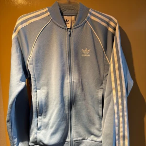 Ljusblå Adidas track jacket - Ljusblå Adidas track jacket med klassiska vita ränder längs ärmarna och broderad logga på bröstet. Jackan har dragkedja framtill, ribbade muddar och ståkrage. Tillverkad i en mjuk blandning av polyester och bomull.