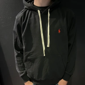 Svart hoodie från Polo Ralph Lauren - Svart hoodie från Polo Ralph Lauren med vit dragsko och röd broderad logga på bröstet. Klassisk känguruficka framtill och mjukt material. Perfekt för en avslappnad stil.
