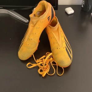 Gula fotbollsskor från Adidas - Säljer ett par gula Adidas fotbollsskor med svarta ränder och orange sula. Skorna har snörning och är designade för att ge bra grepp på planen. Perfekta för dig som vill sticka ut på matchen!