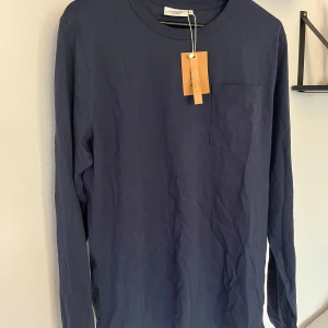 Mörkblå långärmad tröja från Dobber - Säljer en mörkblå långärmad tröja från Dobber i storlek L. Tröjan har en enkel design med rund halsringning och en bröstficka. Perfekt för en stilren och avslappnad look. Aldrig använd, prislapp är kvar.