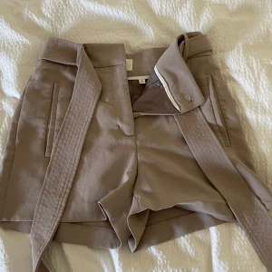 Beige shorts med bälte - Stilrena beige shorts med tillhörande bälte och hög midja. Shortsen har raka ben, diskreta fickor fram och bak samt hällor för bältet. Perfekta för en dressad look och tillverkade i ett mjukt tyg.