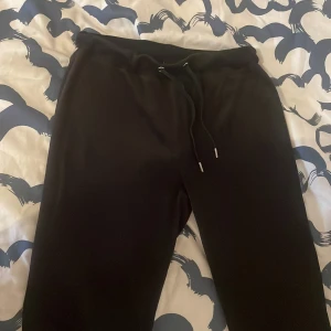 Svarta velour mjukis från Gina Tricot - Svarta mjukisbyxor från Gina Tricot med dragsko i midjan och två bakfickor. Byxorna har en avslappnad passform och är perfekta för en chill stil. Tillverkade i mjukt material för extra komfort.