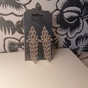 Glittriga örhängen från Red Carpet Collection - Säljer ett par långa, glittriga örhängen i silverfärg från Red Carpet Collection by madlady. Örhängena har ett detaljerat, bladliknande mönster och är perfekta för att ge en extra touch till din outfit. För stora för min smak men perfekt på en ute kväll men kompisar eller ens kille helt nya 
