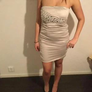 Champagnefärgad axelbandslös klänning med paljettdetaljer - Tror är oanvänd storlek passar 34-38. Säljer en snygg axelbandslös kortklänning i champagnefärg med glittriga paljetter upptill. Klänningen har en figurnära passform och är tillverkad i ett glansigt material som ger en lyxig känsla. Perfekt för speciella tillfällen!se gärna mina andra saker jag säljer över har 30 klänningar skicka pm tack .se bilder på min fb sida som heter allt ska bort med röd bakgrund.