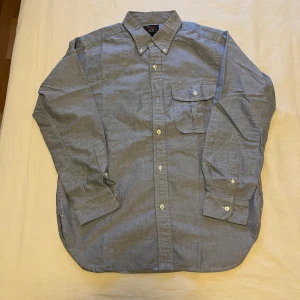 skjorta från Woolrich - Klassisk blå/grå skjorta från Woolrich med knappar hela vägen, bröstficka och button-down krage. Skjortan är långärmad och tillverkad i ett tunt bomullstyg som passar perfekt till många tillfällen. I nästintill perfekt skick. Lite skada vi kragen men inget som syns