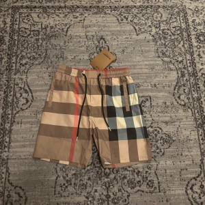Rutig badshorts från Burberry - Snygga badshorts från Burberry med klassiskt rutigt mönster i beige, svart, vitt och rött. Shortsen har elastisk midja med snörning och bakficka. Perfekta för sommaren och tillverkade i ett lätt syntetmaterial.