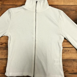 Vit ribbad jacka med dragkedja - Säljer en vit ribbad kofta med hög krage och hel dragkedja framtill. Kofta har lång ärm och en enkel, stilren design som passar perfekt till vardags. Materialet är mjukt och stretchigt för extra komfort. Koftan är one size, dock skulle den passa bra för dig som har storlek Xxs, xs, s och m