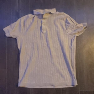 Ljusgrå ribbad pikétröja - Säljer en ljusgrå pikétröja med ribbad struktur och klassisk krage. Tröjan har korta ärmar och knäppning med tre knappar framtill. Perfekt för en avslappnad och stilren look. Storlek S