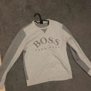 Hugo Boss sweatshirt  - Säljer en grå sweatshirt från Hugo Boss med stora BOSS-trycket framtill. Tröjan har långa ärmar i två olika grå nyanser och klassisk rund halsringning. Perfekt för en avslappnad stil.