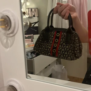 Mönstrad handväska från Tommy Hilfiger - Säljer en snygg handväska från Tommy Hilfiger med svart och beige mönster, svarta handtag och detaljer i rött och grönt med guldfärgade knappar. Väskan har dragkedja och är perfekt för dig som vill ha en stilren accessoar.